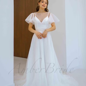 May include: A white wedding dress with a sheer bodice and a flowing skirt. The dress has short, ruffled sleeves and a scoop neckline. The model is wearing the dress in a studio setting.