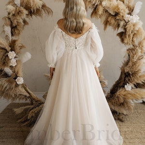 May include: A white off-the-shoulder wedding dress with long, puffy sleeves and a lace bodice. The dress has a flowing skirt and is photographed from the back.