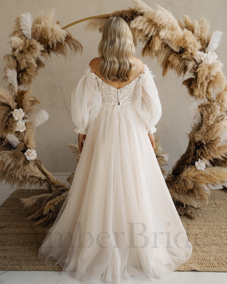 May include: A white off-the-shoulder wedding dress with long, puffy sleeves and a lace bodice. The dress has a flowing skirt and is photographed from the back.