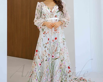 May include: A white, floor-length gown with a floral embroidery design. The dress features a V-neckline, long sleeves, and a flowing skirt with a train. The embroidery includes red, pink, and purple flowers with green stems.