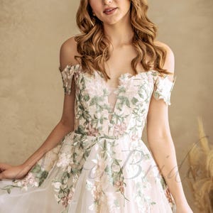 May include: A white off-the-shoulder wedding dress with floral embroidery in shades of green and pink. The dress has a fitted bodice and a flowing skirt.