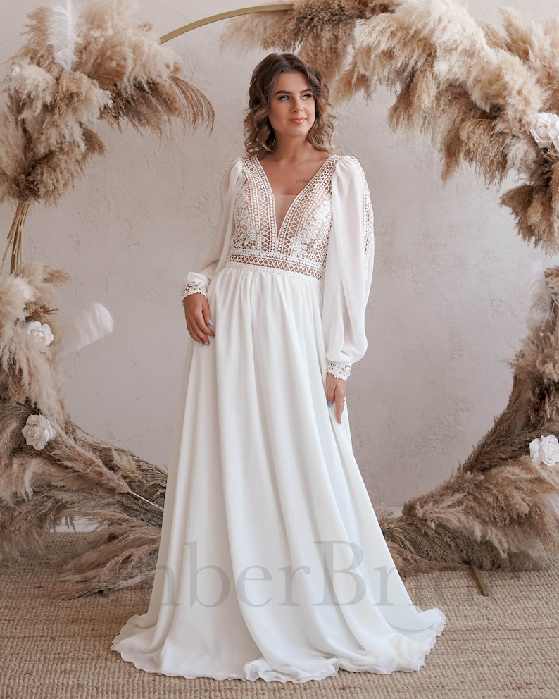 May include: A white, long-sleeved, lace wedding dress with a V-neckline and a flowing skirt. The dress has a fitted bodice and a sheer overlay.