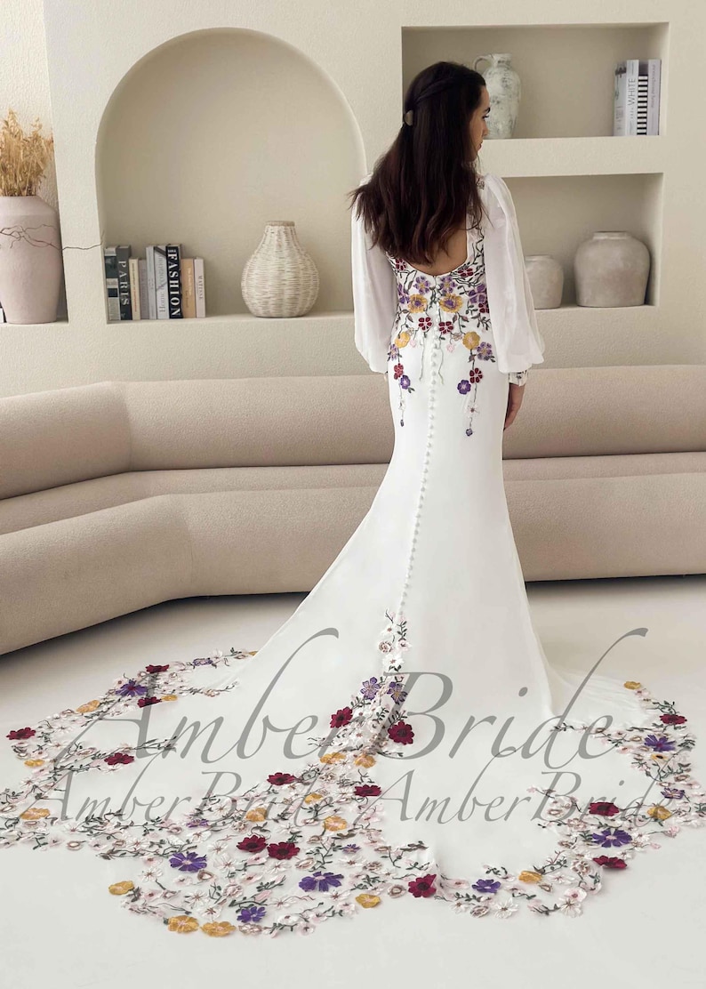 May include: White wedding dress with floral embroidery in red, purple, and yellow. The dress features long, sheer sleeves and a long train with matching floral detailing. The back of the dress has a row of buttons.