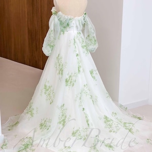May include: A white off-the-shoulder gown with a green floral print. The gown has a full skirt and a fitted bodice. The gown is made of a sheer fabric and has a ruffled neckline.