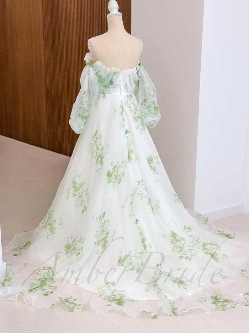 May include: A white off-the-shoulder gown with a green floral print. The gown has a full skirt and a fitted bodice. The gown is made of a sheer fabric and has a ruffled neckline.