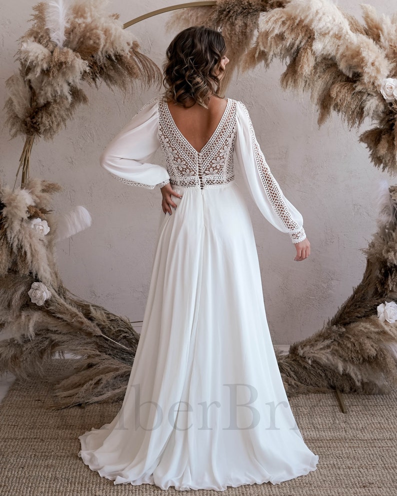 May include: A white long-sleeved wedding dress with a lace bodice and a flowing skirt. The dress has a deep V-neckline and a button-up back.