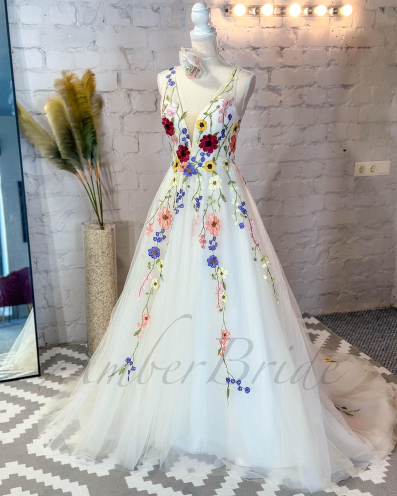 May include: A white tulle gown with a V-neckline and floral embroidery. The embroidery features a variety of colors, including pink, blue, yellow, and red.