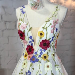 May include: A white tulle dress with a deep V-neckline and colorful floral embroidery. The dress has a fitted bodice and a flowing skirt.