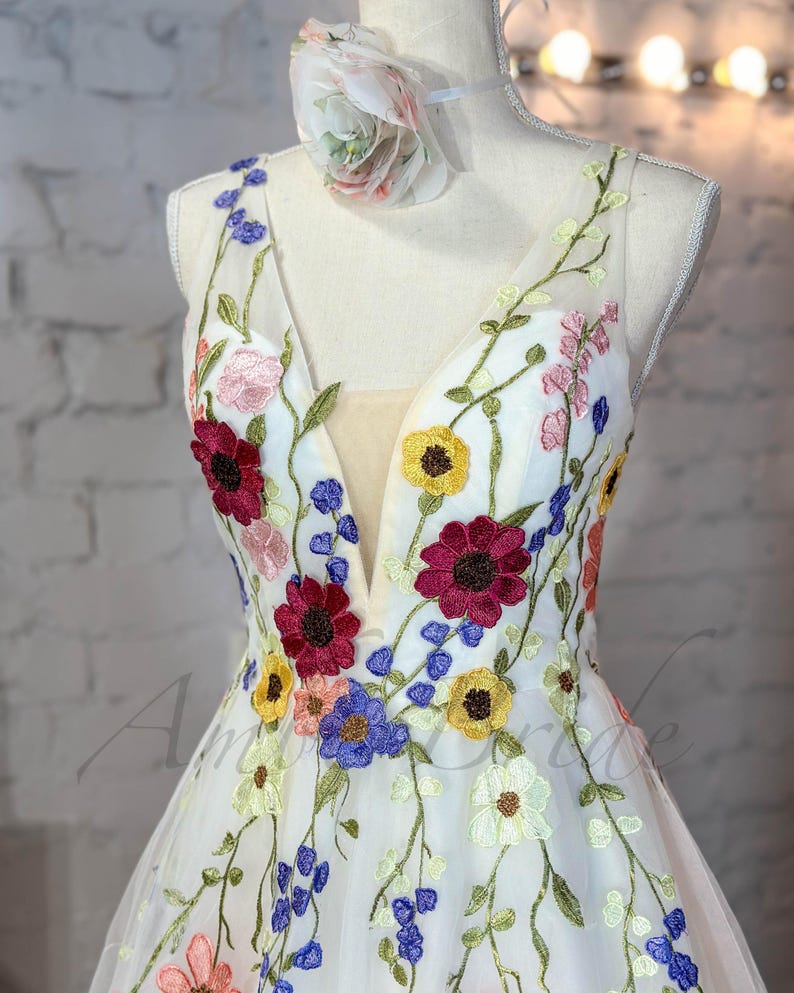May include: A white tulle dress with a deep V-neckline and colorful floral embroidery. The dress has a fitted bodice and a flowing skirt.