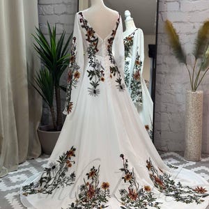 May include: A white maxi dress with long sleeves and a V-neckline. The dress is adorned with a floral print in shades of green, brown, and orange.