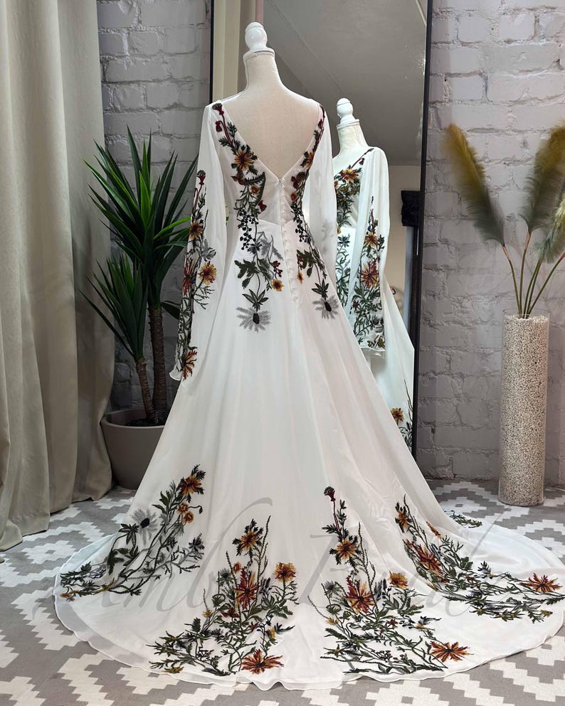 May include: A white maxi dress with long sleeves and a V-neckline. The dress is adorned with a floral print in shades of green, brown, and orange.
