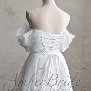 May include: A white wedding dress with a corset back and off-the-shoulder sleeves. The dress is displayed on a mannequin, showcasing the delicate fabric and design. The text "Amber Bride" is visible.