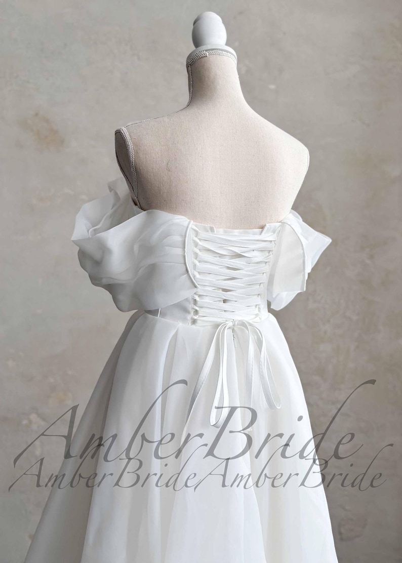 May include: A white wedding dress with a corset back and off-the-shoulder sleeves. The dress is displayed on a mannequin, showcasing the delicate fabric and design. The text "Amber Bride" is visible.