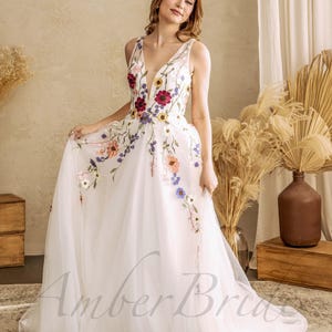 May include: A white tulle wedding dress with a plunging neckline and colorful floral embroidery. The dress has a full skirt and a long train.