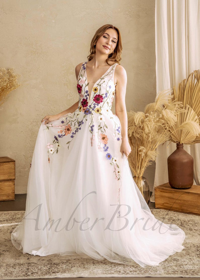 May include: A white tulle wedding dress with a plunging neckline and colorful floral embroidery. The dress has a full skirt and a long train.