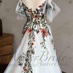 May include: White tulle wedding dress with a floral embroidery design. The dress has a V-neckline, a fitted bodice, and a flowing skirt. The embroidery features a variety of flowers in shades of red, orange, yellow, and green.