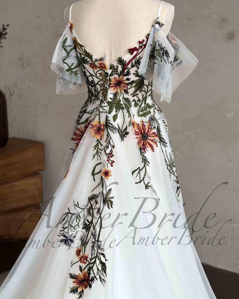 May include: White tulle wedding dress with a floral embroidery design. The dress has a V-neckline, a fitted bodice, and a flowing skirt. The embroidery features a variety of flowers in shades of red, orange, yellow, and green.
