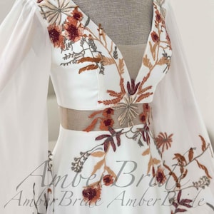 May include: White wedding dress with a V-neckline and long, sheer sleeves. The bodice and sleeves are adorned with intricate floral embroidery in shades of rust, brown, and green. A sheer panel accents the waist. The dress is displayed on a mannequin.