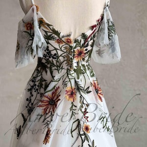 May include: A white tulle dress with floral embroidery. The dress has a V-neckline, off-the-shoulder straps, and a flowing skirt. The embroidery features a variety of flowers in shades of red, orange, yellow, and green.