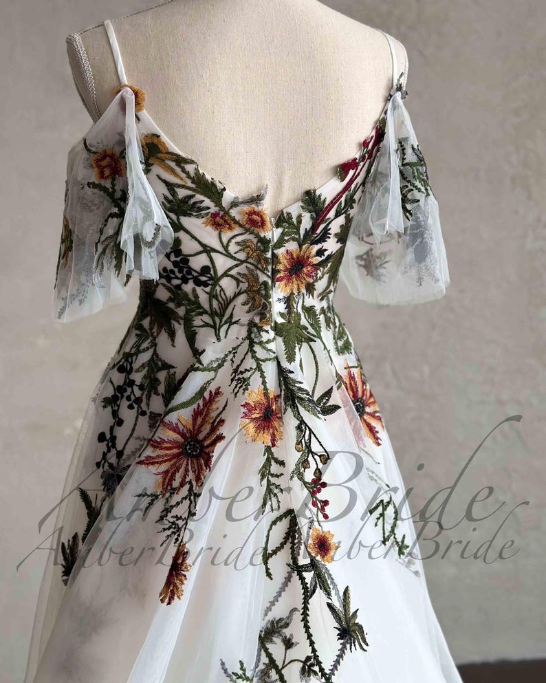 May include: A white tulle dress with floral embroidery. The dress has a V-neckline, off-the-shoulder straps, and a flowing skirt. The embroidery features a variety of flowers in shades of red, orange, yellow, and green.