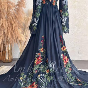 May include: A flowing black dress with long sleeves and a deep V-neck. The dress is adorned with colorful floral embroidery in red, yellow, and white. The dress has a long train and is made of a sheer fabric.