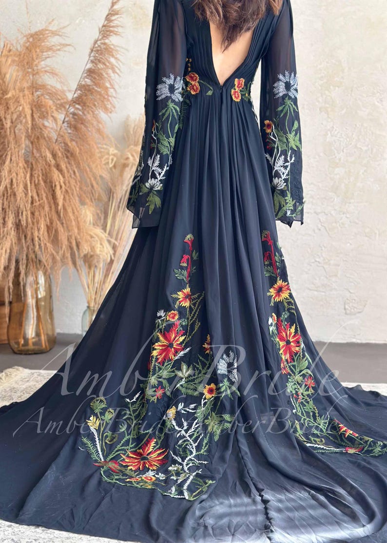 May include: A flowing black dress with long sleeves and a deep V-neck. The dress is adorned with colorful floral embroidery in red, yellow, and white. The dress has a long train and is made of a sheer fabric.
