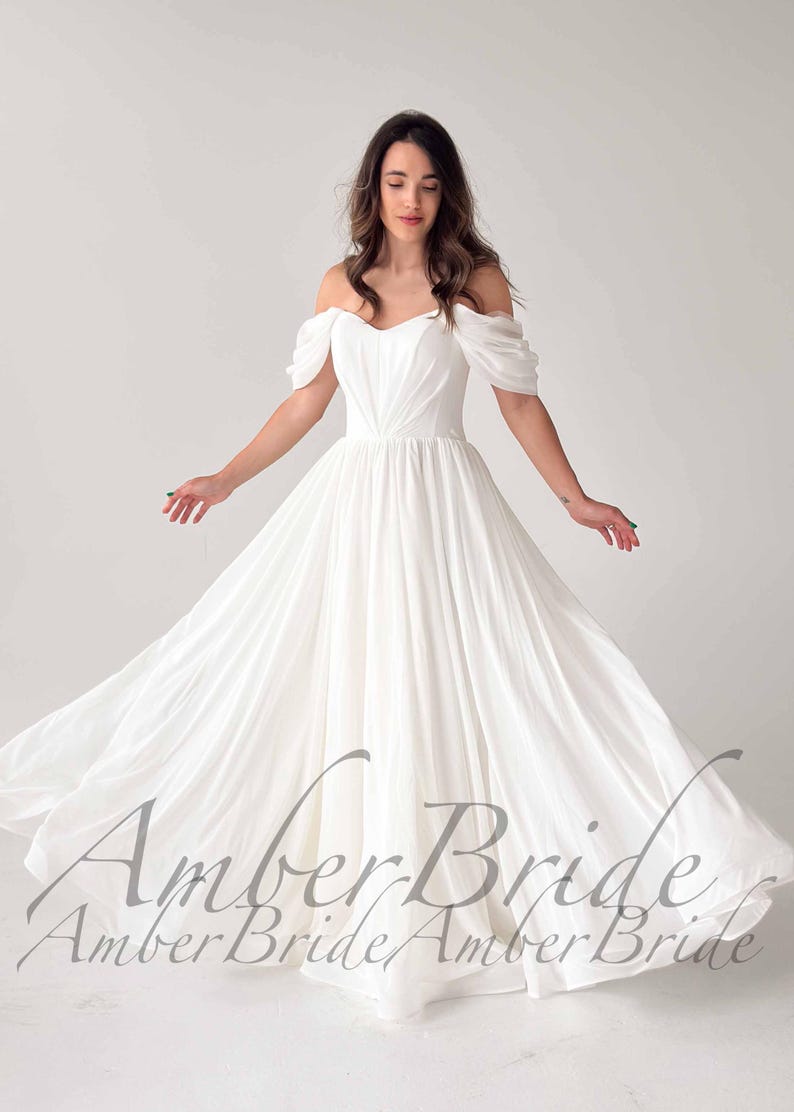 May include: A white off-the-shoulder wedding dress with a flowing skirt. The dress features a sweetheart neckline and short, draped sleeves. The text "Amber Bride" is printed across the bottom of the image.