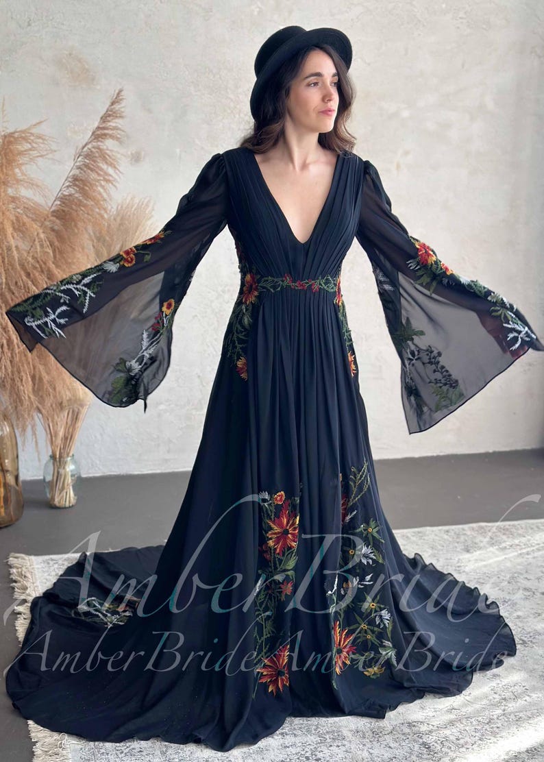 May include: A black, floor-length dress with long, sheer sleeves and a plunging neckline. The dress is adorned with colorful floral embroidery along the sleeves, waist, and skirt. The dress has a flowing silhouette and a train.