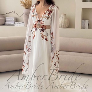 May include: A white wedding dress with a plunging neckline and sheer sleeves. The dress features floral embroidery in shades of brown and orange. The waistline is accentuated with a matching floral design. The dress has a flowing skirt and a train.
