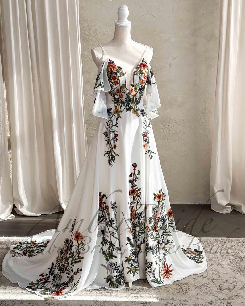May include: A white wedding dress with floral embroidery. The dress features a plunging neckline, spaghetti straps, and a flowing skirt. The floral design includes red, orange, and green accents. The dress is displayed on a mannequin.