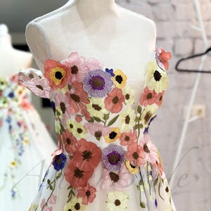 May include: A white off-the-shoulder dress with a floral embroidery design. The dress features a variety of colorful flowers, including pink, yellow, blue, and purple.