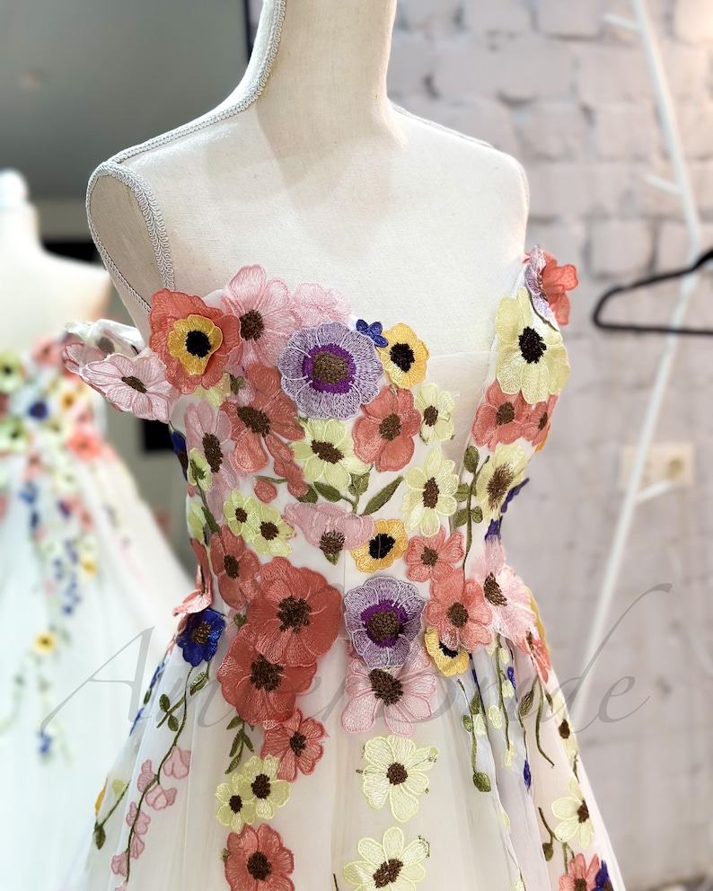 May include: A white off-the-shoulder dress with a floral embroidery design. The dress features a variety of colorful flowers, including pink, yellow, blue, and purple.