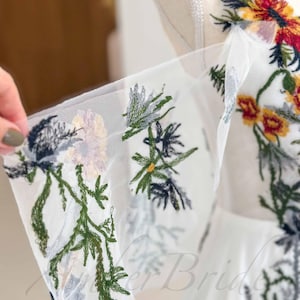 May include: White tulle fabric with embroidered floral designs in shades of green, blue, yellow, and red.