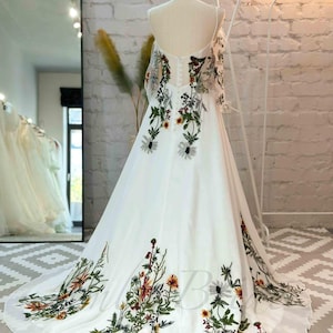 May include: A white wedding dress with a floral pattern. The dress has a sweetheart neckline and a flowing skirt. The floral pattern is in shades of green, yellow, and orange.