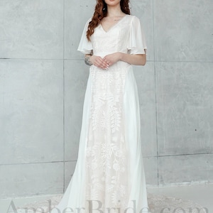 May include: A white wedding dress with a V-neckline and a lace overlay. The dress has a flowing skirt and a train. The model is wearing the dress with a simple hairstyle and makeup. The dress is photographed against a white background. AmberBride.com