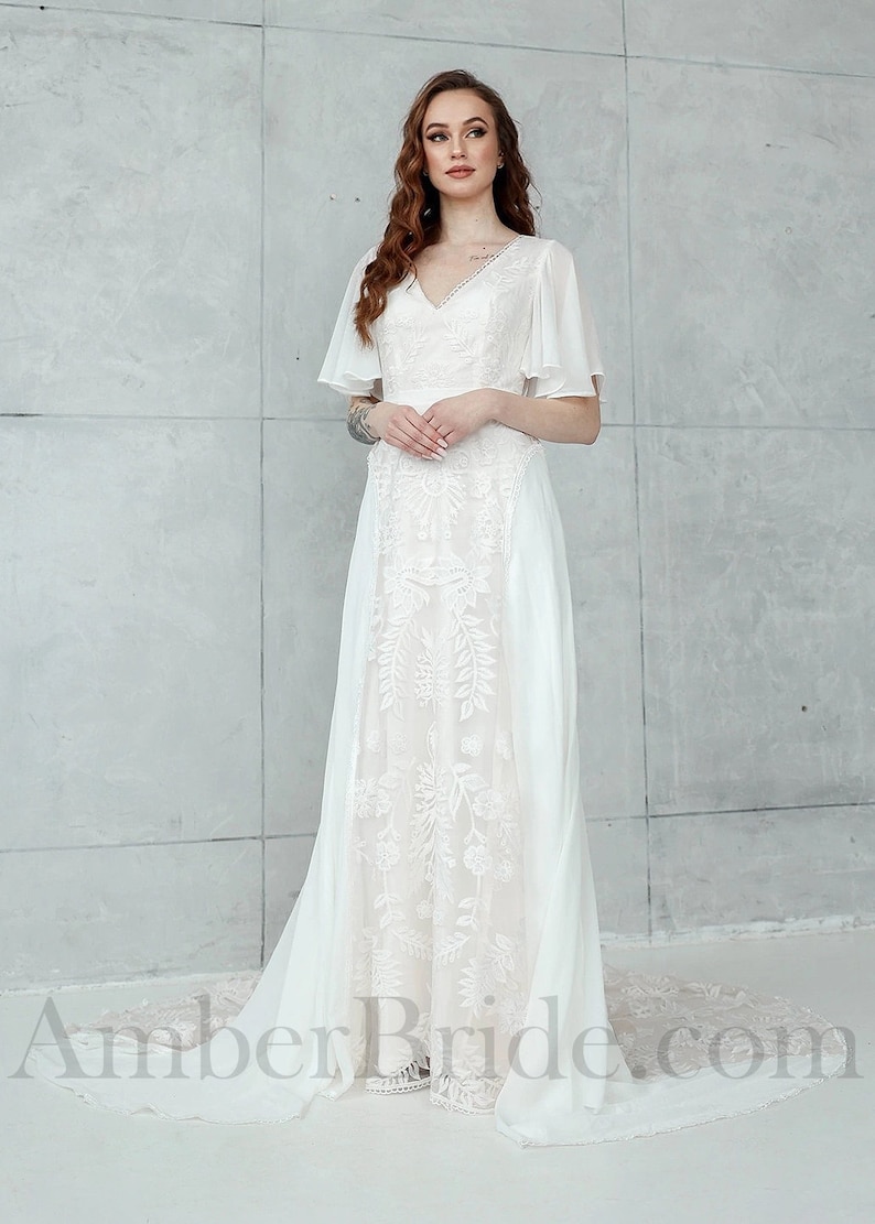 May include: A white wedding dress with a V-neckline and a lace overlay. The dress has a flowing skirt and a train. The model is wearing the dress with a simple hairstyle and makeup. The dress is photographed against a white background. AmberBride.com