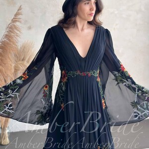 May include: Black chiffon dress with a plunging neckline and long, flowing sleeves. The dress features floral embroidery in red, yellow, and green along the waist and sleeves. A black hat completes the look. The text "Amber Bride" is visible on the dress.