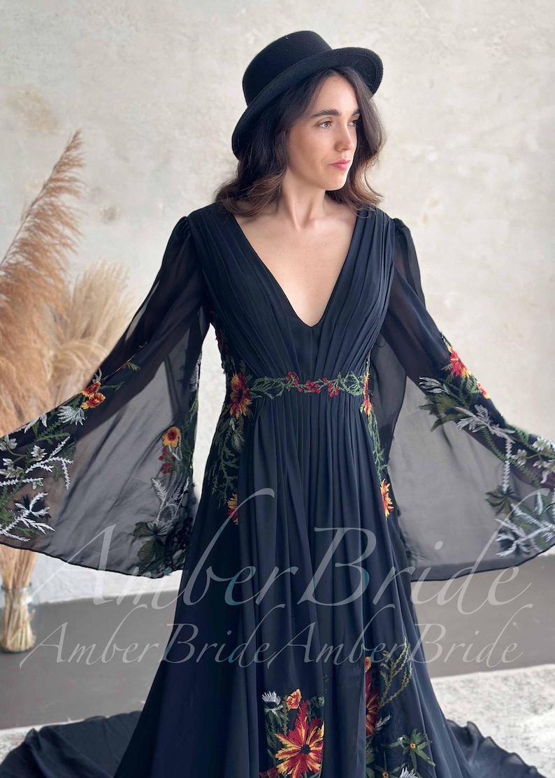 May include: Black chiffon dress with a plunging neckline and long, flowing sleeves. The dress features floral embroidery in red, yellow, and green along the waist and sleeves. A black hat completes the look. The text "Amber Bride" is visible on the dress.