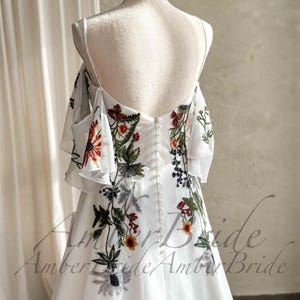 May include: White wedding dress with floral embroidery. The dress features a low back, spaghetti straps, and off-the-shoulder sleeves. The embroidery includes red, orange, green, and black floral designs. The dress has a button-up back and a flowing skirt.