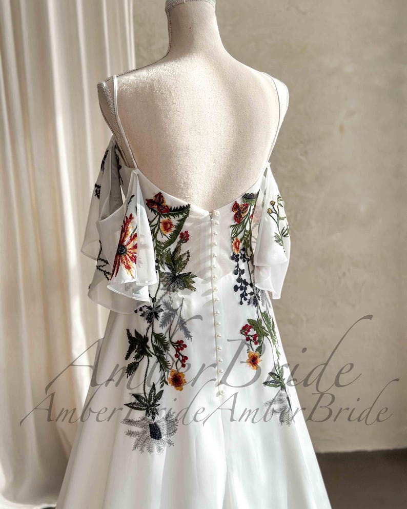 May include: White wedding dress with floral embroidery. The dress features a low back, spaghetti straps, and off-the-shoulder sleeves. The embroidery includes red, orange, green, and black floral designs. The dress has a button-up back and a flowing skirt.