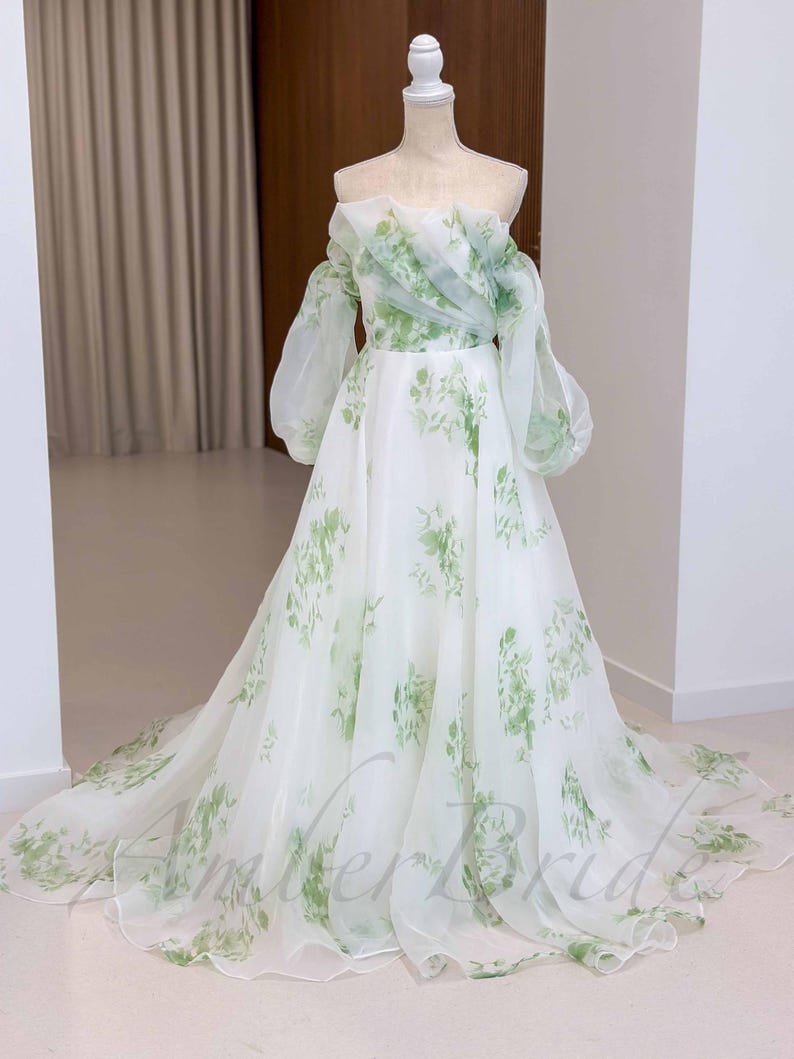May include: A white off-the-shoulder gown with a floral print in shades of green. The gown has a full skirt and long, puffy sleeves.