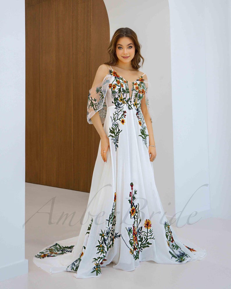May include: A white maxi dress with a floral embroidery design. The dress has a V-neckline, a fitted bodice, and a flowing skirt. The embroidery features a variety of flowers in shades of red, orange, yellow, and green.