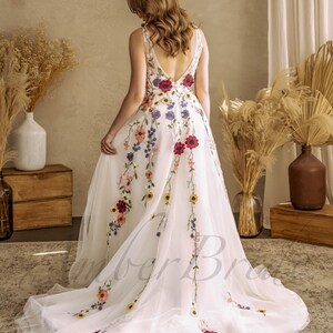 May include: A white tulle wedding dress with a floral embroidery design. The dress has a V-neckline and a low back. The embroidery features a variety of colorful flowers, including red, pink, yellow, and blue.
