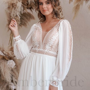 May include: A white, long-sleeved wedding dress with a plunging neckline and delicate lace detailing. The dress has a flowing skirt and sheer sleeves.