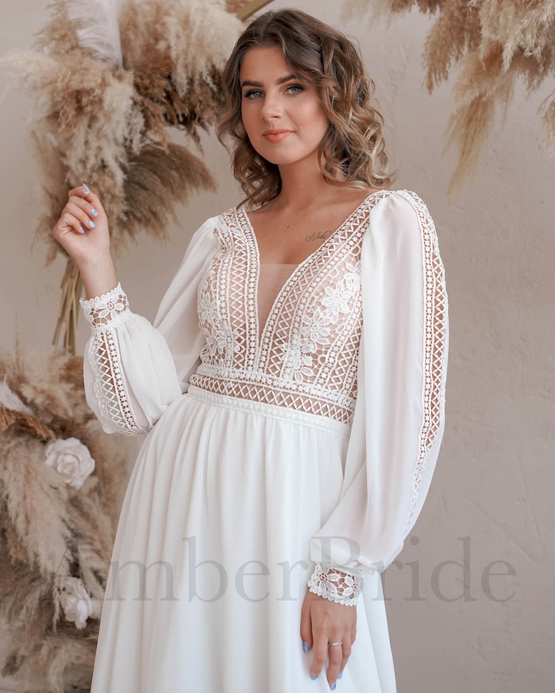 May include: A white, long-sleeved wedding dress with a plunging neckline and delicate lace detailing. The dress has a flowing skirt and sheer sleeves.