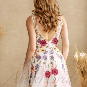 May include: A white tulle wedding dress with a deep V-back and colorful floral embroidery. The dress has a flowing skirt and a fitted bodice.
