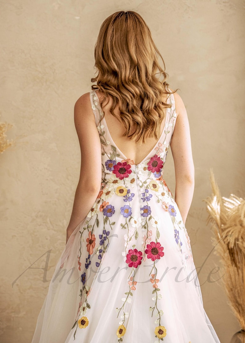 May include: A white tulle wedding dress with a deep V-back and colorful floral embroidery. The dress has a flowing skirt and a fitted bodice.