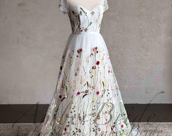 Puede incluir: Un vestido de tul blanco con un corpiño transparente y mangas cortas. El vestido está bordado con flores de colores y vegetación.
