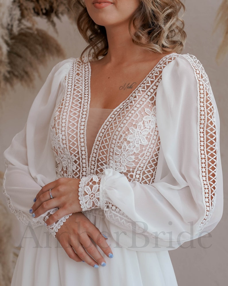 May include: A white bridal gown with long sleeves and a deep V-neckline. The sleeves and neckline are trimmed with delicate lace. The gown is made of a soft, flowing fabric.