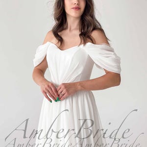May include: A white off-the-shoulder wedding dress with a flowing skirt. The dress features draped sleeves and a sweetheart neckline. The text "Amber Bride" is visible in a script font at the bottom of the image.